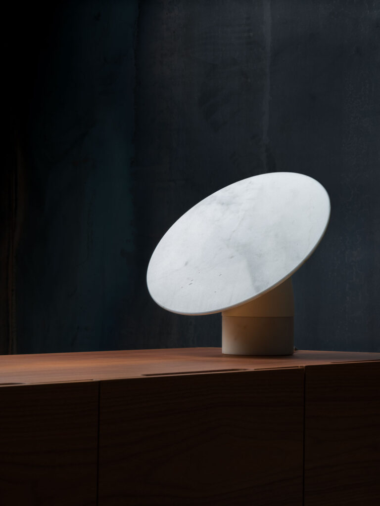 fotografia still-life di lampada di design siciliano in marmo bianco di carrara poggiato su mobile di legno su fondo scuro.