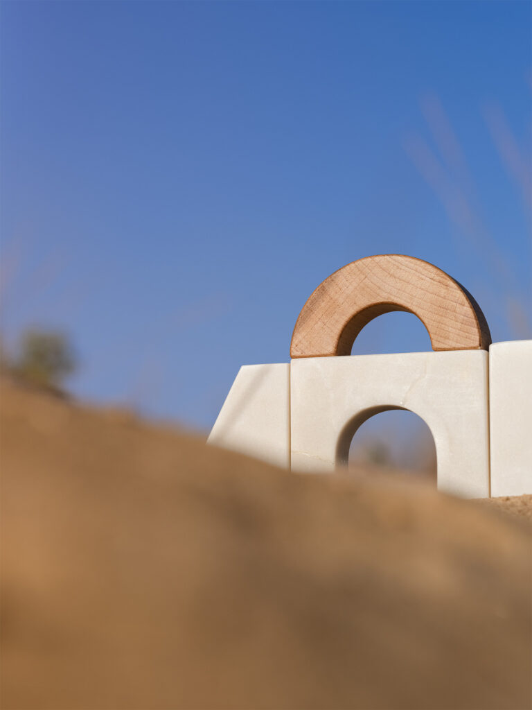 dettaglio fotografia still-life oggetto di design siciliano con pezzi di marmo bianco e legno sulla sabbia con sfondo cielo azzurro.